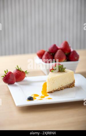 Une tranche de cheesecake crémeux garnie d’une fraise fraîche, servie sur une assiette blanche avec un filet de sauce aux fruits. Un bol de fraises fraîches Banque D'Images