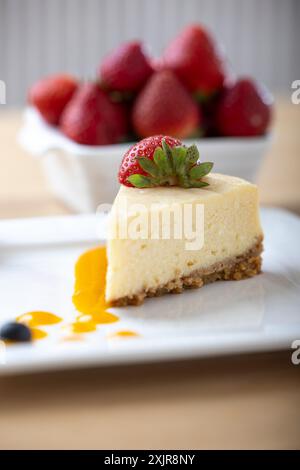 Une tranche de cheesecake crémeux garnie d’une fraise fraîche, servie sur une assiette blanche avec un filet de sauce aux fruits. Un bol de fraises fraîches Banque D'Images