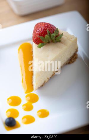 Une tranche de cheesecake crémeux garnie d’une fraise fraîche, servie sur une assiette blanche avec un filet de sauce aux fruits. Un bol de fraises fraîches Banque D'Images