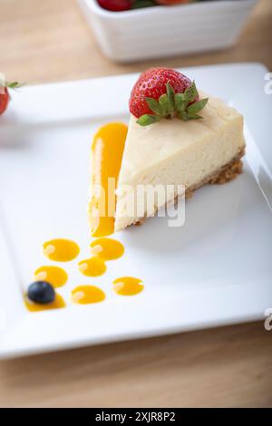 Une tranche de cheesecake crémeux garnie d’une fraise fraîche, servie sur une assiette blanche avec un filet de sauce aux fruits. Un bol de fraises fraîches Banque D'Images