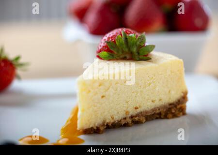 Une tranche de cheesecake crémeux garnie d’une fraise fraîche, servie sur une assiette blanche avec un filet de sauce aux fruits. Un bol de fraises fraîches Banque D'Images