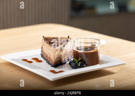 Tranche décadente de cheesecake au chocolat arrosée de sauce au chocolat, servie sur une assiette carrée blanche. Accompagné d'une petite tasse de gan au chocolat riche Banque D'Images