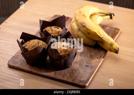 Muffins aux pépites de chocolat à la banane fraîchement cuits enveloppés dans du papier parchemin brun, placés sur une planche à découper en bois à côté d'un bouquet de bananes mûres. Banque D'Images