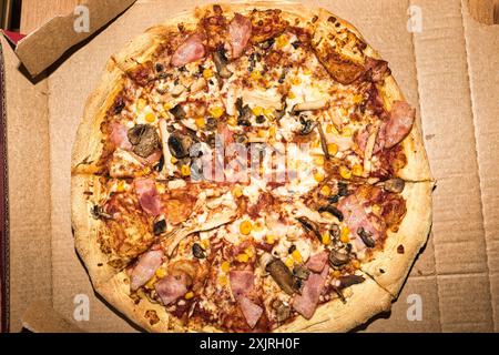 Gros plan d'une pizza avec diverses garnitures, y compris du jambon, des champignons, du maïs et du fromage, placée dans une boîte en carton ouverte. Banque D'Images
