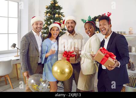 Jeunes amis souriants joyeux multiethniques dans des chapeaux de Père Noël dans le salon célébrant Noël. Banque D'Images