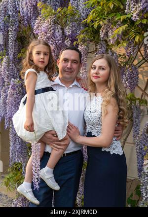 Papa tient doucement une fille mignonne dans ses bras et embrasse maman. Maman et fille sont habillées de belles robes, papa est habillé dans une manche blanche. bac floral Banque D'Images