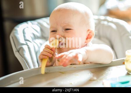 Bébé fille de 6 mois à la maison en chaise haute manger de la nourriture savoureuse à la maison. Bébé drôle avec le visage de purée de brocoli sain souillé tient la cuillère dans les mains Banque D'Images