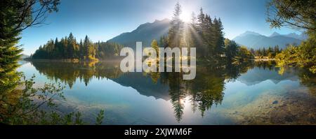 Paysage de lac peint avec des montagnes et des arbres reflétés dans l'eau. Un paysage panoramique idyllique avec de beaux rayons de soleil Banque D'Images