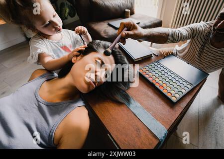 La jeune fille et son père se maquillent sur leur mère, qui est allongée sur une table les yeux fermés, et qui semble fatiguée Banque D'Images