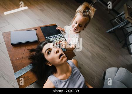 Petite fille applique soigneusement le maquillage à sa mère, qui est allongée sur une table avec les yeux grands ouverts, profitant d'un moment ludique à la maison Banque D'Images