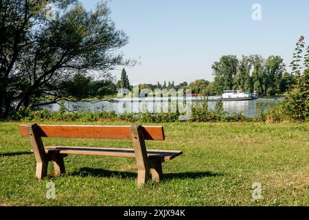 Un banc en bois avec un siège en bois brun et des pieds en béton est placé sur une pelouse herbeuse verte. Le banc fait face à une rivière avec une barge plus grande qui passe Banque D'Images