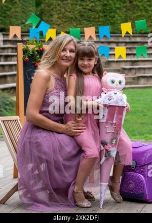 Maman embrasse fermement sa petite fille, qui est allée en 1re année. La fille tient une corne d'école. Sur le fond d'un conseil scolaire et multicolore Banque D'Images