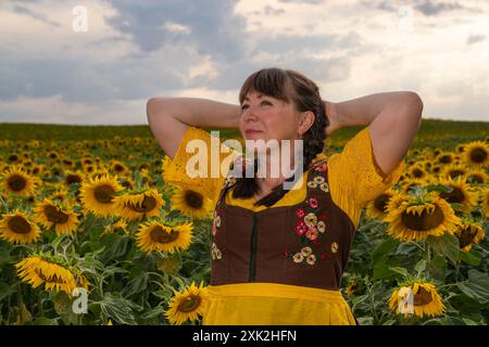 Une femme se tient dans un champ avec des tournesols jaunes. Ses cheveux sont tressés, elle porte une blouse jaune, une robe de soleil brune et un tablier jaune. Banque D'Images