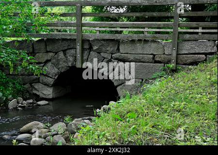 Un pont de pierre rustique avec une balustrade en bois enjambe un petit ruisseau entouré de verdure luxuriante et de rochers. L'arche du pont et le cadre naturel Banque D'Images