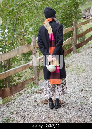 Une femme sans visage vêtue d'une tenue d'automne élégante tient une théière, prête pour une heure de thé en plein air confortable, alors qu'elle marche sur un chemin de gravier pittoresque entouré Banque D'Images