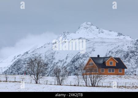 Une maison orange vibrante se démarque du paysage enneigé et des montagnes escarpées des Lofoten, en Norvège, dépeignant une scène hivernale tranquille Banque D'Images