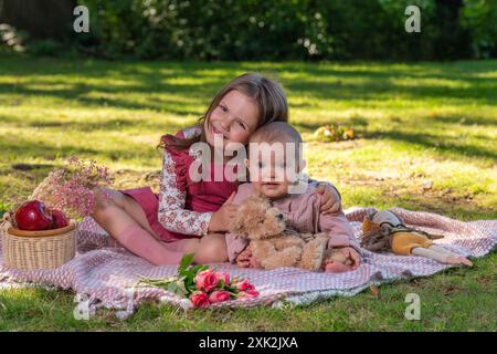 Portrait de jolies filles de 5 ans et 1 an. Deux sœurs en pique-nique. Ils ont l'air amusants devant la caméra. Il y a un ours en peluche à proximité et une application rouge Banque D'Images