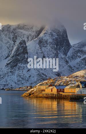 Une vue d'hiver époustouflante sur les Lofoten, avec des maisons en bois vibrantes perchées sur pilotis au bord des eaux calmes et glacées, en toile de fond par mountai enneigé Banque D'Images
