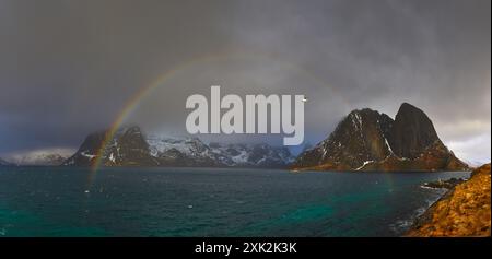 Une vue panoramique époustouflante capture un arc-en-ciel vibrant qui surplombe les sommets glacés et accidentés des Lofoten, avec de sombres nuages orageux et une mer tranquille en toile de fond Banque D'Images