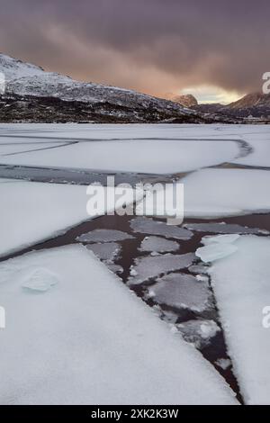 Scène hivernale éclatante dans les Lofoten avec glace fragmentée au-dessus d'un lac tranquille avec une montagne spectaculaire en toile de fond sous un ciel sombre Banque D'Images