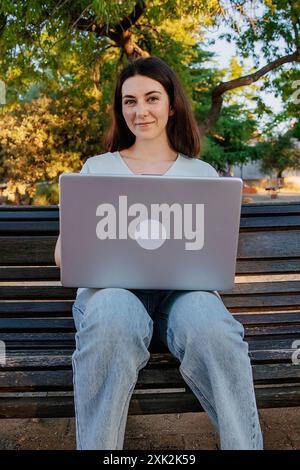 Jeune femme regardant la caméra tout en travaillant sur son ordinateur portable, assise sur un banc de parc entouré d'arbres à la lumière du soleil. Banque D'Images