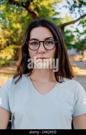 Portrait d'une jeune femme caucasienne portant des lunettes et un t-shirt bleu clair, regardant directement la caméra avec une expression neutre, debout dans un Banque D'Images