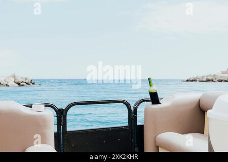 Une vue sereine depuis un bateau montrant une bouteille de vin placée sur une balustrade, surplombant une mer calme et la côte rocheuse sous un ciel clair parfait pour les profondeurs Banque D'Images