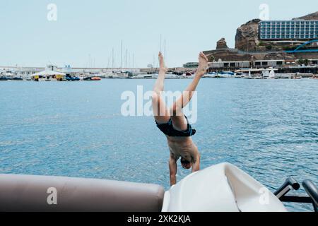 Un homme effectue un saut du bord d'un bateau dans les eaux cristallines de la mer, avec un port animé et un bâtiment au sommet d'une colline dans le backgroun Banque D'Images