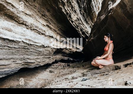 Une jeune femme dans un bikini rouge vif trouve la tranquillité tout en méditant dans une grotte rocheuse isolée, incarnant une retraite d'été paisible loin du h. Banque D'Images