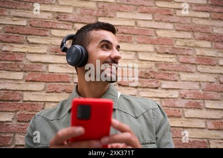 Gros plan jeune homme souriant avec des écouteurs tout en tenant un téléphone portable rouge avec fond de mur de briques Banque D'Images
