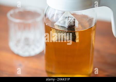 Un pot en verre rempli de thé infusé, avec un sachet de thé infusé à l'intérieur. Le pot est placé sur une surface en bois, avec un gobelet en verre transparent à l'arrière Banque D'Images