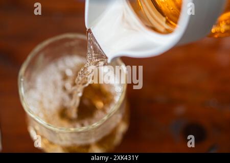 Gros plan d'un verre rempli d'un liquide brun doré provenant d'un pichet blanc. L'arrière-plan est flou, soulignant l'action de versement et Banque D'Images