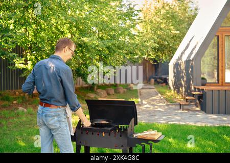 Homme d'âge moyen met une poêle de cuisson avec steak sur le barbecue tout en faisant de la nourriture à l'arrière-cour Banque D'Images