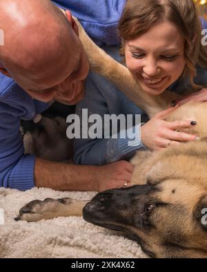 La famille aime être avec le chien. Un homme et une femme sont couchés sur le sol, avec un grand berger allemand couché à proximité. Banque D'Images