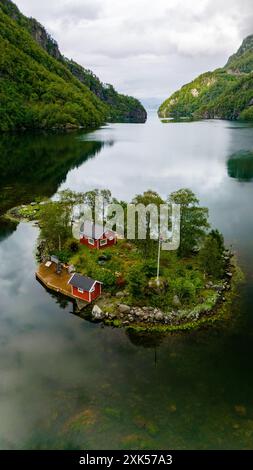 Une cabane rouge nichée sur une petite île dans un fjord entouré de montagnes verdoyantes et d'eau calme. Lovrafjorden, Norvège Banque D'Images