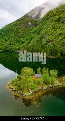 Une scène idyllique d'une cabane rouge nichée sur une petite île dans un fjord norvégien, entourée de verdure luxuriante et de montagnes couvertes de brume. Lovrafjorden, Norvège Banque D'Images