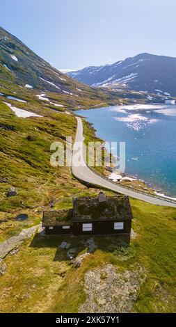 Une route pittoresque serpente à travers un paysage montagneux, au bord d'un lac immaculé avec des sommets enneigés au loin. Une petite cabine se trouve nichée sur la colline. Langvatnet, Geiranger, Norvège Banque D'Images