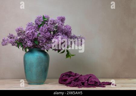 Un grand bouquet de lilas dans un vase turquoise sur un fond clair, avec un châle violet foncé couché à côté. Nature morte avec des fleurs. Carte postale, con Banque D'Images