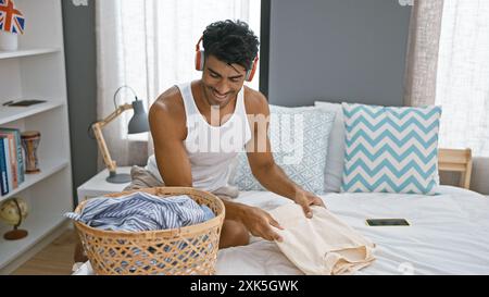 Jeune homme hispanique souriant pliant le linge dans une chambre lumineuse tout en écoutant de la musique avec des écouteurs rouges Banque D'Images