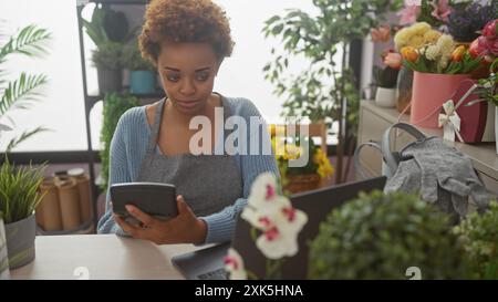 Femme africaine utilisant la calculatrice dans un magasin de fleurs rempli de bouquets colorés et de plantes vertes. Banque D'Images