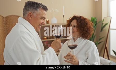 Couple d'âge moyen dégustant du vin ensemble dans un centre de bien-être spa portant des peignoirs blancs Banque D'Images
