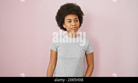 Femme afro-américaine aux cheveux bouclés posant dans une chemise grise décontractée sur un fond isolé rose. Banque D'Images