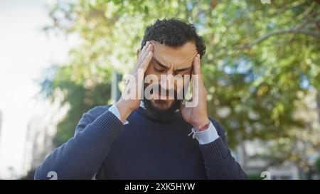 Homme d'âge moyen stressé avec la barbe touchant les tempes dans un parc vert montrant mal de tête ou frustration à l'extérieur. Banque D'Images