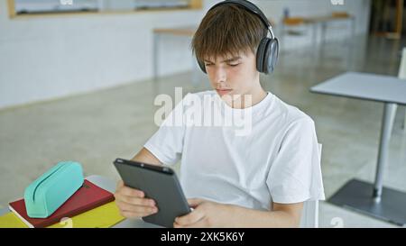 Un jeune homme concentré étudiant avec une tablette dans une bibliothèque universitaire tout en portant des écouteurs. Banque D'Images