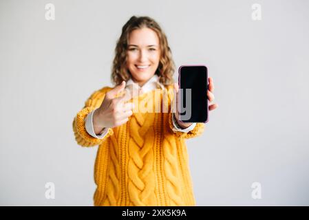 Femme heureuse pointant vers l'écran de téléphone mobile, jeune femme souriante pointant vers l'écran de téléphone mobile, présentant une nouvelle application. Tenir un smartphone avec un Banque D'Images