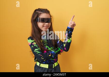 Adorable fille hispanique pointe joyeusement sur le côté, son visage illuminé d'un sourire confiant alors qu'elle navigue dans un monde de réalité virtuelle à travers son regard Banque D'Images