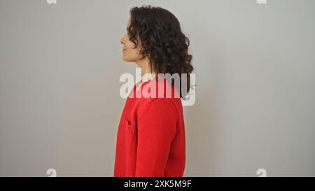 Attrayante jeune femme hispanique brune dans un pull rouge pose contre un mur de fond blanc isolé. Banque D'Images