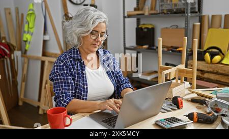Une femme mature dans une chemise à carreaux concentrée sur un ordinateur portable dans son atelier de travail du bois encombré. Banque D'Images
