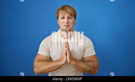 Un jeune homme blond avec une barbe, portant un t-shirt décontracté, se tient debout sur un fond bleu extérieur avec ses mains serrées dans un geste de plaidoyer. Banque D'Images