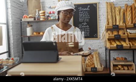 Jeune, africain, femme, in, boulangerie, porter, tablier, debout, à, comptoir avec, pain, fond. Banque D'Images